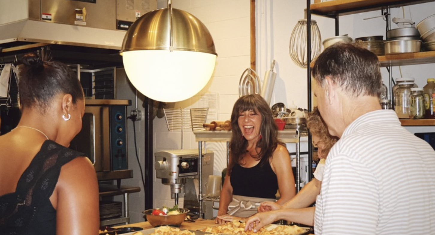 A diverse group of people stand at a kitchen table in a restaurant, smiling and laughing over a spread of food